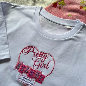 May include: A light blue t-shirt with a crew neck. The shirt features an embroidered design of a heart with the words "Pretty Girl" in cursive and "AVENUE" in block letters. Below the heart is the text "where your dreams come true."