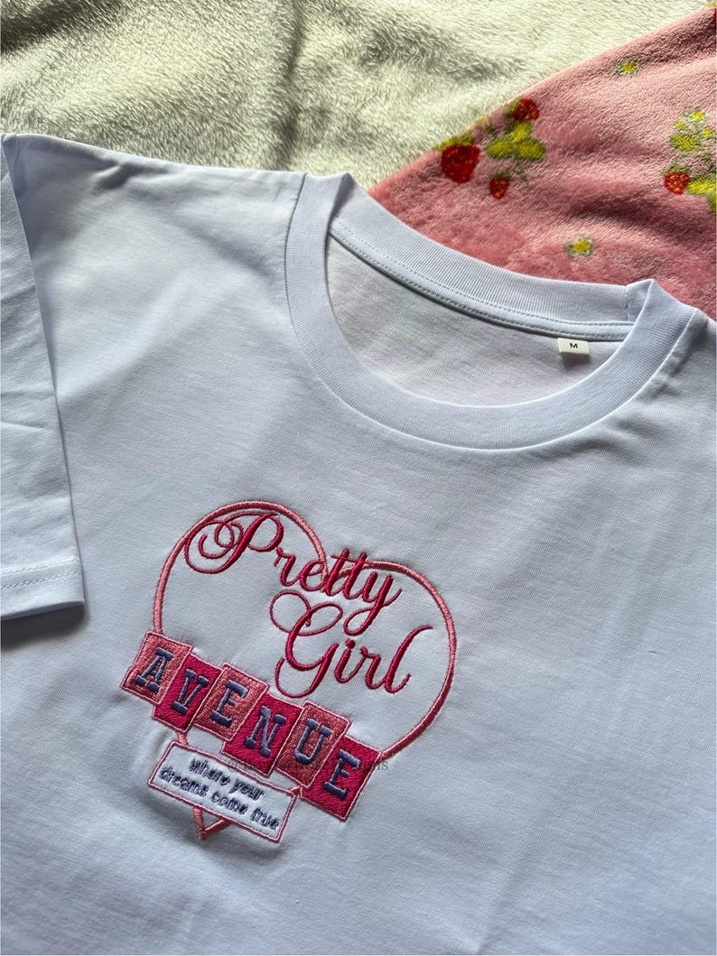 May include: A light blue t-shirt with a crew neck. The shirt features an embroidered design of a heart with the words "Pretty Girl" in cursive and "AVENUE" in block letters. Below the heart is the text "where your dreams come true."