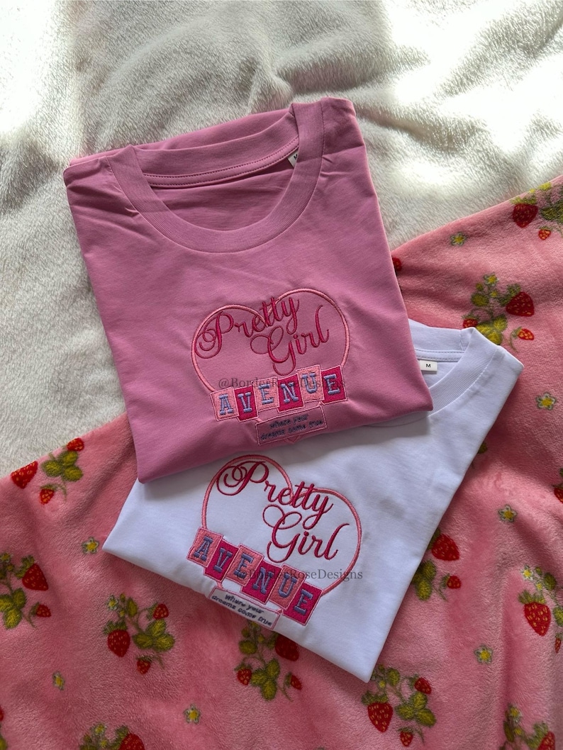 May include: Two t-shirts, one pink and one white, with embroidered designs. The pink shirt has a "Pretty Girl" heart design with "Avenue" and text. The white shirt has the same design. Both shirts are on a pink strawberry-patterned blanket.