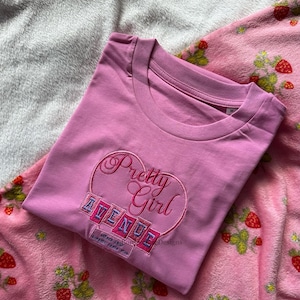 May include: A pink t-shirt with the embroidered text "Pretty Girl Avenue" in a heart-shaped design. The shirt is folded and placed on a pink strawberry-patterned blanket.
