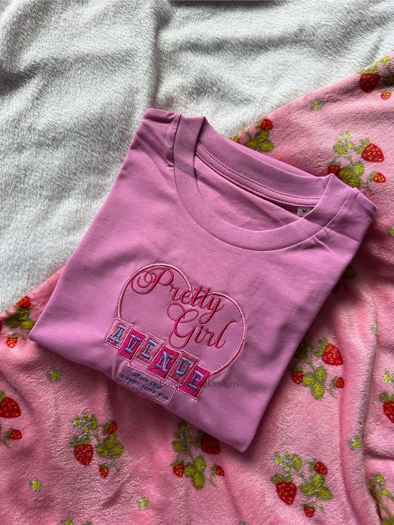 May include: A pink t-shirt with the embroidered text "Pretty Girl Avenue" in a heart-shaped design. The shirt is folded and placed on a pink strawberry-patterned blanket.