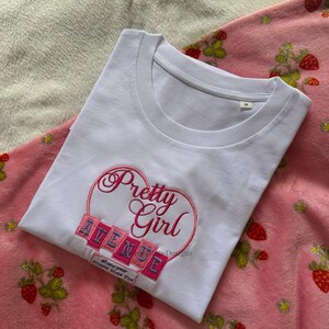 May include: White t-shirt with the embroidered text "Pretty Girl Avenue" inside a pink heart. The shirt is on a pink strawberry-patterned blanket. The text "Where your dreams come true" is below the word "Avenue".