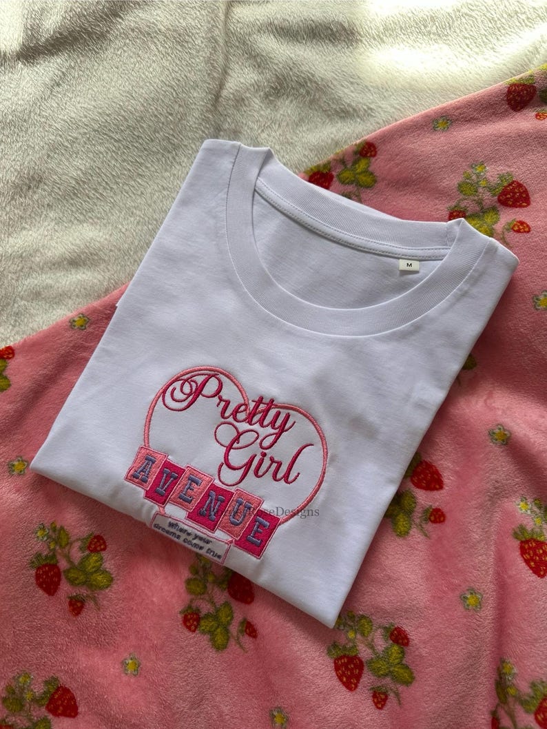 May include: White t-shirt with the embroidered text "Pretty Girl Avenue" inside a pink heart. The shirt is on a pink strawberry-patterned blanket. The text "Where your dreams come true" is below the word "Avenue".