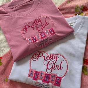May include: Two t-shirts, one pink and one white, with the embroidered text "Pretty Girl Avenue" in a heart-shaped design. The pink shirt is on top of the white shirt. The text "where your dreams come true" is also embroidered.