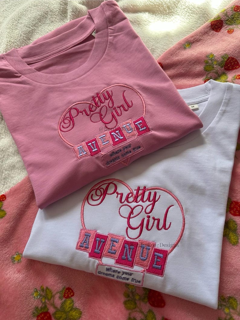 May include: Two t-shirts, one pink and one white, with the embroidered text "Pretty Girl Avenue" in a heart-shaped design. The pink shirt is on top of the white shirt. The text "where your dreams come true" is also embroidered.