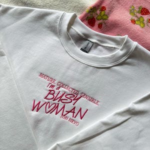 Pode incluir: Um moletom branco com gola redonda com o texto bordado em rosa: "MATURE, COLLECTED, SENSIBLE, I'M A BUSY WOMAN (OH HEY)". O moletom &eacute; feito de um material macio. Um cobertor rosa com estampas de morango &eacute; vis&iacute;vel no fundo.