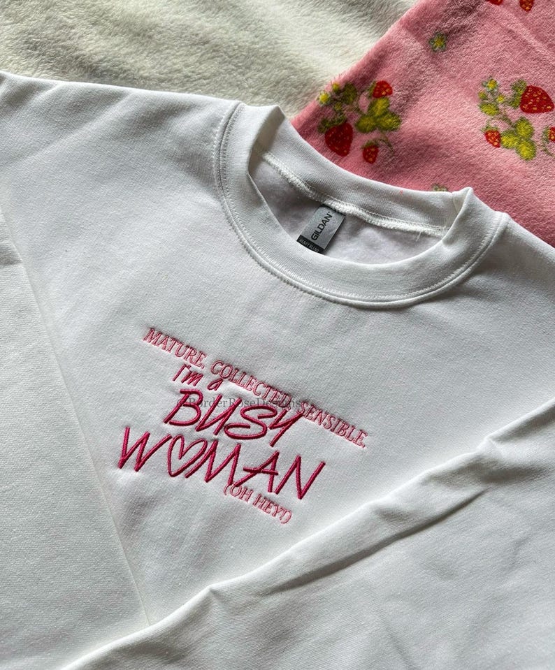 Pode incluir: Um moletom branco com gola redonda com o texto bordado em rosa: "MATURE, COLLECTED, SENSIBLE, I'M A BUSY WOMAN (OH HEY)". O moletom &eacute; feito de um material macio. Um cobertor rosa com estampas de morango &eacute; vis&iacute;vel no fundo.