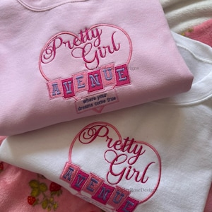 May include: Two sweatshirts, one pink and one white, are displayed. Both feature the embroidered text "Pretty Girl Avenue" in a heart-shaped design. The pink sweatshirt has the phrase "where your dreams come true" embroidered below the main text.