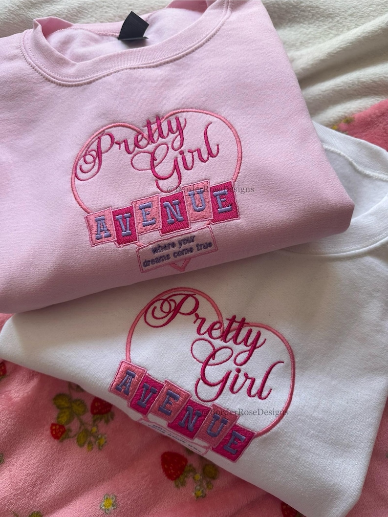 May include: Two sweatshirts, one pink and one white, are displayed. Both feature the embroidered text "Pretty Girl Avenue" in a heart-shaped design. The pink sweatshirt has the phrase "where your dreams come true" embroidered below the main text.