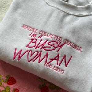 Pode incluir: Camisola branca com texto rosa bordado: &laquo;MATURE, COLLECTED, SENSIBLE, I'M A BUSY WOMAN (OH HEY!)&raquo;. A camisola de gola redonda &eacute; feita de um material macio.