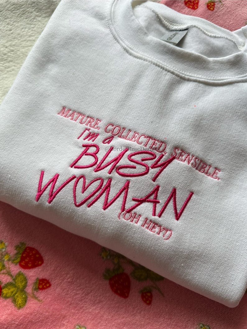 Pode incluir: Camisola branca com texto rosa bordado: &laquo;MATURE, COLLECTED, SENSIBLE, I'M A BUSY WOMAN (OH HEY!)&raquo;. A camisola de gola redonda &eacute; feita de um material macio.