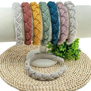 May include: A collection of braided headbands in various colors, including gray, brown, yellow, green, teal, and pink. The headbands are displayed on a white stand, with one headband lying flat below. The headbands have a thick, textured appearance.