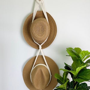 May include: Two brown straw hats hanging on a white macrame hat rack with wooden beads. The rack is attached to a white wall. A green potted plant is in the lower right corner of the image.