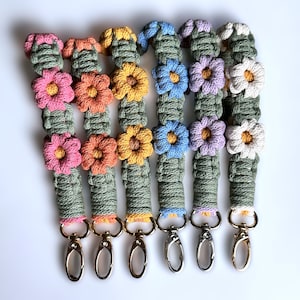 May include: Six colorful crocheted keychains with flower designs and gold metal key rings. The keychains are made with green, pink, orange, yellow, blue, and white yarn.