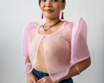 May include: A woman wearing a sheer pink top with puffed sleeves and a black choker necklace. She is also wearing a pair of pink tassel earrings and a gold bracelet. She is smiling and looking at the camera.