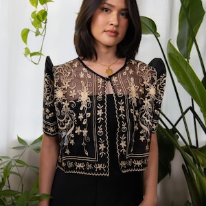 May include: A black bolero jacket with intricate gold floral embroidery. The sheer fabric reveals a glimpse of the outfit beneath. The jacket features short sleeves and a scalloped hemline, creating an elegant and detailed look.