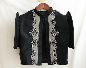 May include: A black, short-sleeved jacket with white floral embroidery. The jacket features a mandarin collar and is displayed on a wooden hanger. The fabric has a sheen. The garment is likely made of a synthetic material.