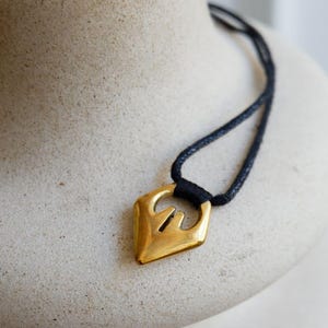 May include: A gold pendant necklace with a geometric design. The pendant is shaped like a diamond with a small hole in the centre. The necklace is made of black cord.