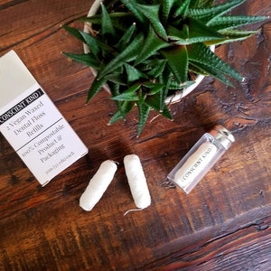 Puede incluir: Una caja blanca con el texto "Consent Kind" y "1 Vegan Waxed Dental Floss Refills" en ella. La caja está junto a dos recambios de hilo dental blancos y un recipiente de vidrio transparente con una tapa de corcho. El recipiente también tiene el texto "Consent Kind" en él. Los artículos están sobre una superficie de madera.