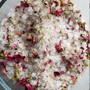May include: A close-up of a bowl filled with white bath salts, rose petals, and dried flowers. The bath salts are a light pink colour and the petals are a deep red.