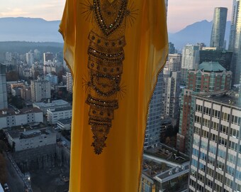 May include: A yellow kaftan with a beaded design hangs in front of a window with a city view.