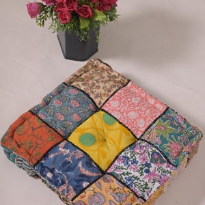 Peut inclure: Coussin carré en patchwork avec une variété de tissus à motifs floraux dans les tons orange, bleu, rose et vert. Le coussin a une bordure noire et un centre jaune. Un vase de roses roses et jaunes est en arrière-plan.