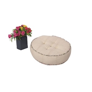 Peut inclure: Un coussin de sol rond crème avec capitonnage et bordure à motifs. Un vase carré noir contient un bouquet de roses roses, jaunes et rouges. Les articles sont posés sur un fond blanc.
