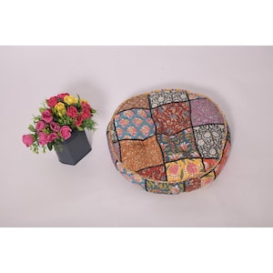 Peut inclure: Un coussin de sol rond en patchwork avec divers motifs floraux et géométriques dans les tons de bleu, marron, rouge et jaune. Un petit bouquet de fleurs roses, jaunes et rouges dans un vase gris foncé est également présent.