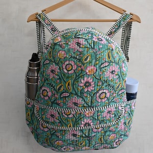 Può includere: Uno zaino trapuntato color verde acqua con un motivo floreale rosa, giallo e verde. Lo zaino ha spallacci a righe bianche e nere e tasche laterali che contengono una bottiglia d'acqua argentata e una bottiglia blu. È appeso a una gruccia di legno.
