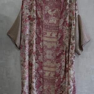 May include: A floral print kimono in shades of pink, cream, and brown hangs on a wooden hanger. The kimono features a mix of floral and geometric patterns, with a solid pink panel down the center. The sleeves have a brown polka dot pattern.