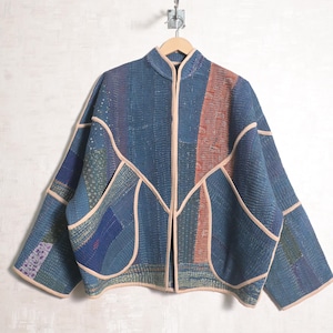 May include: A quilted jacket featuring a patchwork design of various blue fabric panels with visible stitching. The jacket has a mandarin collar, long sleeves, and light beige piping along the front opening, pockets, and hemline edges.