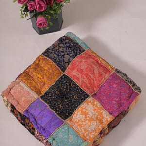 Puede incluir: Cojín cuadrado de patchwork con una variedad de telas estampadas con motivos florales en tonos naranja, morado, azul y rosa. El cojín está acolchado y tiene un borde decorativo. Un pequeño jarrón negro con flores rosas y amarillas está en el fondo.