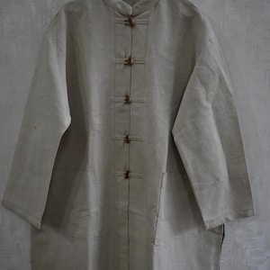Puede incluir: Una camisa larga de lino beige claro con cuello mandarín y cierres de palanca de madera. La camisa tiene dos bolsillos delanteros y está colgada de una percha de madera. La prenda parece tener un corte holgado.