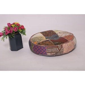 Puede incluir: Cojín de suelo redondo de patchwork con una variedad de cuadrados de tela estampada en tonos marrón, morado y crema. El cojín tiene un borde marrón y está sobre una superficie blanca. Un jarrón de flores rosas y amarillas está a la izquierda.