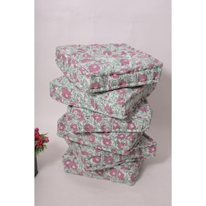 Puede incluir: Una pila de cinco cojines cuadrados para el suelo. Cada cojín presenta un estampado floral con flores rosas y hojas verdes sobre un fondo blanco. Los cojines están acolchados y tienen un aspecto suave y acogedor.