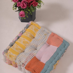 Peut inclure: Coussin carré en patchwork avec des tissus jaunes, oranges, roses et bleus. Le coussin est capitonné. Un arrangement floral dans un récipient noir est visible en arrière-plan. Dimensions : environ 40 cm.