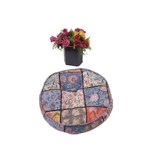 May include: A round patchwork floor cushion with a floral pattern in various colors, including blue, pink, and yellow. Above the cushion is a black square vase filled with pink and yellow roses.