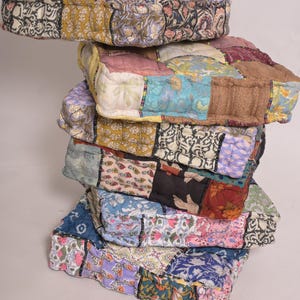 Peut inclure: Une pile de coussins de sol carrés aux motifs patchwork. Les coussins présentent une variété de motifs floraux et géométriques dans les tons de rose, bleu, jaune et marron. Les coussins sont empilés les uns sur les autres.