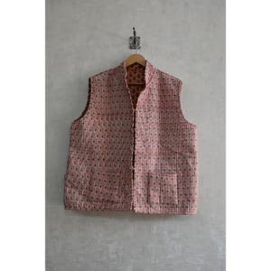以下が含まれることがあります： ピンクとグリーンの模様のキルティングベストが木製のハンガーに掛けられています。ノースリーブのベストは、マンダリンカラー、フロントポケット、ボタン留めが特徴です。生地は質感のある外観をしています。