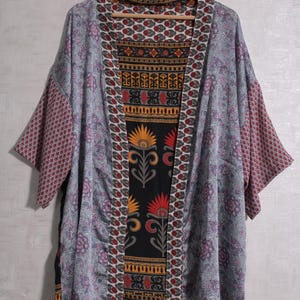 Könnte beinhalten: Eine gemusterte Kimono-Jacke mit einer Mischung aus floralen und geometrischen Designs. Die Jacke hat eine schwarze Tafel mit orangefarbenen und roten Blumenmotiven, die von einem weißen und roten Muster umrandet ist. Die Ärmel und Außenbahnen sind hellgrau mit lila floralen Akzenten.