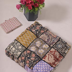 Puede incluir: Cojín cuadrado de patchwork con una variedad de patrones florales y geométricos en tonos amarillos, azules, rojos y negros. También hay una pequeña cartera con estampado floral y un ramo de flores rosas y amarillas en un jarrón negro.