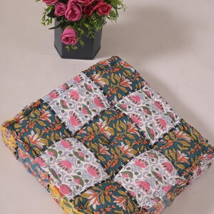 Puede incluir: Cojín cuadrado de patchwork con un estampado floral en verde azulado, rosa y blanco. El cojín está hecho de múltiples cuadrados de tela cosidos. Un jarrón hexagonal negro con rosas rosas y amarillas se encuentra en el fondo.