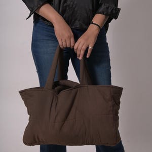 May include: A brown quilted tote bag with two handles. The bag is held by a person wearing blue jeans and a black shirt. The tote bag has a rectangular shape and a padded design.