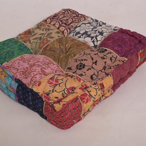 May include: Square patchwork cushion with a variety of fabric patterns and colors. The cushion features a mix of floral, paisley, and geometric designs in shades of red, green, blue, and gold. The edges are a deep burgundy.