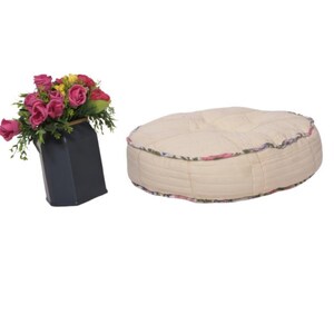 Puede incluir: Un jarrón negro contiene un ramo de rosas rosas y amarillas. Junto a él, un cojín de suelo redondo, color crema, con un ribete floral. El cojín tiene un diseño acolchado.