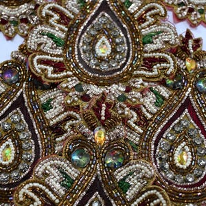 Könnte beinhalten: Nahaufnahme eines Stoffes mit aufwendiger Perlenstickerei und Verzierungen. Das Design zeigt Tropfen- und Blumenmuster in Gold, Burgunderrot, Weiß und Grün. Schimmernde, irisierende Edelsteine sind im Muster verstreut.