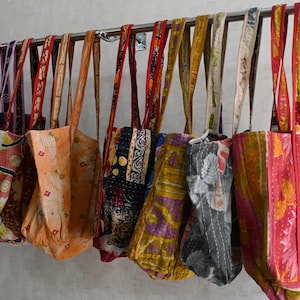 May include: A collection of colorful tote bags hanging on a metal bar. The bags feature various patterns and colors, including floral, geometric, and patchwork designs. The straps are made of matching or contrasting fabric.
