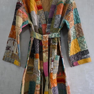 May include: A patchwork robe with a belt, featuring a variety of fabric patterns and colors including yellow, orange, green, and pink. The robe is hanging on a wooden hanger against a light gray wall.