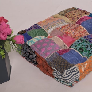 Puede incluir: Un cojín de suelo cuadrado de patchwork con una variedad de patrones de tela coloridos. El cojín está junto a un jarrón gris oscuro lleno de rosas rosas y amarillas. Un artículo decorativo para la decoración del hogar.