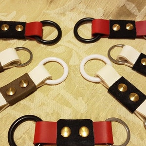 May include: Five different leather keyrings with two metal rings. Each keyring has a black leather strap with two gold-coloured metal rivets. The straps are in different colours: red, white, and brown.